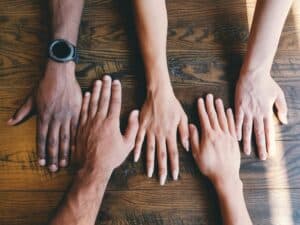 Five hands laying palm side down on a wooden table - people coming together to support a loved one with mental health issues